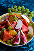 High angle of crop ceramic bowl of slices of different fruits placed on blue surface near blurred grapes