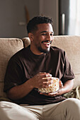 Fröhlicher, lockig behaarter, ethnischer Afroamerikaner in Freizeitkleidung, der auf einem bequemen Sofa sitzt, wegschaut und leckeres Popcorn isst, während er am Wochenende fernsieht