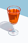 A clear, stemmed glass filled with an amber-colored liquid against a white surface with a blue background, casting a sharp shadow.