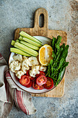 Top view of vibrant dish of raw vegetables with sliced cucumber, halved cherry tomatoes, cauliflower pieces, a lemon slice, and arugula on a cutting board