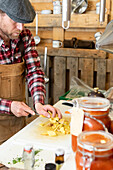 Crop focused chef in apron and beret standing in a rustic kitchen attentively dicing potatoes on a white cutting board with a background of hanging utensils and jars of homemade preserves