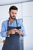 Adult bearded male chef in blue shirt and apron looking at screen of mobile phone while standing in modern kitchen and browsing for text messages