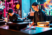 Beautiful focused young chef with braided hair cooking with male coworker in modern kitchen at illuminated sushi bar