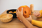 Anonymous women sitting at table and carving with tool scary teeth on orange pumpkin and preparing for Halloween decoration day