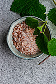 Veganes Kochgeschirr mit einer Schale rosa Himalaya-Salz, garniert mit einem frischen Nesselblatt auf grauem Hintergrund