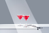 Glasses filled with red pomegranate cocktails placed on a reflective surface under a beam of soft light, creating symmetrical shadows