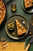 Top view of bread piece with green olives placed with complete oven baked Italian focaccia preparation in pan on dining table with knife and fork