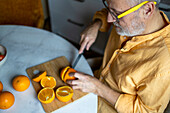 From above crop senior man with glasses and in casual clothes cutting fresh organic oranges with knife on table while sitting in kitchen at home