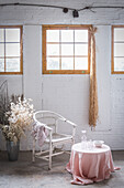 Innenansicht eines hellen Raums mit weißen Backsteinwänden und Holzfenstern, der mit einem Stuhl und einem mit Stoff bezogenen Tisch neben dekorativen Blumen in einer Vase eingerichtet ist