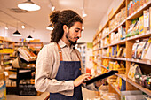 Portrait side view of serious ethnic male worker with long curly hair in casual clothes standing near shelves and working on tablet in modern lighted grocery shop
