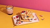 From above tasty sweet waffles topped with berries fruits sauce and cream served on pink tray on colorful table background near glass with hot milk beverage in light kitchen