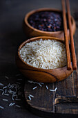 High angle of Raw wild black rice and peeled white rice in the bowls with chopsticks placed on cutting board on wooden table