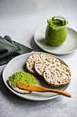High angle of rice bread on a plate accompanied by a vibrant green spinach pesto pasta-sauce in a glass jar, set against a blurred backdrop near napkin