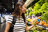 Side view of young female customer in casual clothes with shopping basket choosing fresh carrots while standing near shelves in supermarket