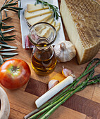 A variety of fresh ingredients including artisan cheese, olive oil, tomatoes, and asparagus laid out on a wooden cutting board.