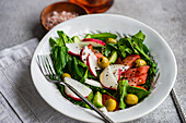 Freshly prepared vegetable salad featuring organic spinach leaves, ripe tomatoes, crisp radishes, and green olives beautifully presented in a white bowl with a fork set against a muted backdrop