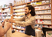Cheerful bearded male in casual clothes with apron standing at table by drug shelves while dispensing drugs in bag to crop customer in pharmaceutical shop