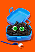 Horror lunch with black pasta and eyes in lunchbox placed on orange background near fork with eye