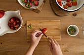From above of anonymous person with knife slicing strawberries on wooden board and during strawberry jam preparation indoors