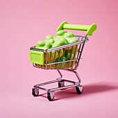 Composition of miniature shopping trolley with green mockup product placed on pink background
