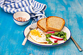 Nahaufnahme einer gesunden Lunch-Schale mit Brotscheiben, Spiegelei, frischer Gurke, Radieschen und Tomate auf einer blauen Fläche neben einem Tuch, daneben eine kleine Schale mit rosa Salz