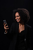 Positive Hispanic young female in outfit smiling and browsing mobile phone while standing on black background