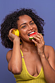 Optimistic African American female model with headphones smiling and looking at camera while biting juicy delicious watermelon in summer