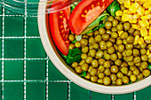 Top view of crop salad bowl with slices of tomato, spinach leaves, corn kernels and peas placed on green surface