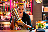 Confident young beautiful barmaid with concentration pouring alcohol in glass from bottle while preparing beverage at modern bistro