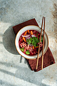 Schale mit Rote-Bete-Suppe mit Zwiebeln, Koriander und Nudeln im asiatischen Stil, serviert auf einer Schale und Stäbchen auf einer Serviette vor einer grauen Fläche im Tageslicht (Draufsicht)