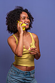 Smiling Hispanic female in casual clothes with curly black hair talking on mobile phone while looking away against blue background