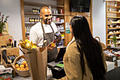 Cheerful African American male shopkeeper in apron standing at counter with barcode scanner and speaking to unrecognizable female customer with long hair