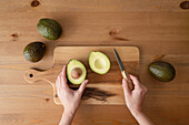 Top view of crop anonymous person with knife cutting ripe avocado on cutting board over wooden table indoor