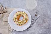 From above top view of ring-shaped choux pastry topped with white icing and sprinkled with chopped pistachios, presented on a white plate with a fork and glass, placed on a beige placemat over a textured grey surface