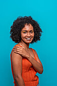 Side view of friendly African American female fashion model in casual clothes with curly hair smiling while standing against blue background