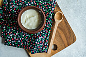 Top view of traditional serving of Georgian sour yogurt known as Matsoni in clay pot near wooden spoon on colorful napkin on wooden tray against blurred background