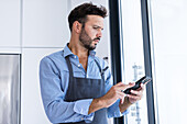 Side view of adult bearded male chef in blue shirt and apron looking at screen of mobile phone while standing near glass window in sunny daylight and browsing for text messages