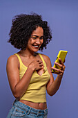Cheerful Hispanic female with Afro hairstyle in yellow top browsing smartphone while standing on blue background