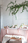 Elegant table served with plates and flowers near yummy cake on pink tablecloth placed near bench covered with cloth against potted plant and white brick wall