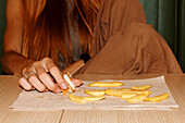Crop anonymous woman with red hair sitting at table and enjoy eating hamburger and french fries in restaurant
