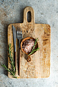 From above rustic kitchen setup on wooden cutting board with a fork, knife, a sprinkle of pink Himalayan salt, fresh rosemary sprigs, laid out on a textured concrete background