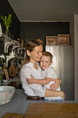 Loving Caucasian mid adult mother hugging his son cute adorable son while standing and together in kitchen during weekend