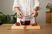 Crop anonymous female in white gown standing at wooden table with jar of fresh strawberry ham