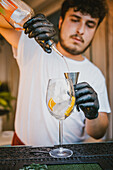 Concentrated young professional barman pouring orange drink in jigger while preparing refreshing cocktail and standing at counter in bar