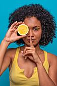Portrait of young Hispanic female with curly black hair covering eye with lemon slice and gesturing silence against blue background