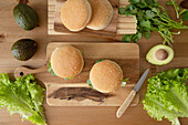 Top view of fresh delicious hamburger with buns and ripe avocado lettuce and cilantro with knife placed on wooden cutting board on table