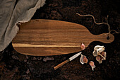 Draufsicht auf ein ovales Schneidebrett aus Holz mit Messer und Knoblauch auf einer rauen Oberfläche