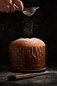 A hand gently dusts powdered sugar onto a panettone, capturing the snow-like sprinkles mid-air against a dark setting