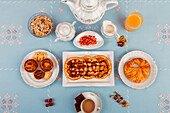 Draufsicht auf ein kontinentales Frühstück mit Gebäck, Müsli, frischem Obst und Getränken, ausgelegt auf einer blauen Fläche