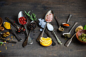 Top view of collection of spoons of various sizes filled with a variety of spices and seeds, such as sunflower seeds, anise star, rosemary, mint, lemon slices, ginger, and assorted peppers, neatly displayed on a dark wooden surface for a rustic appeal.